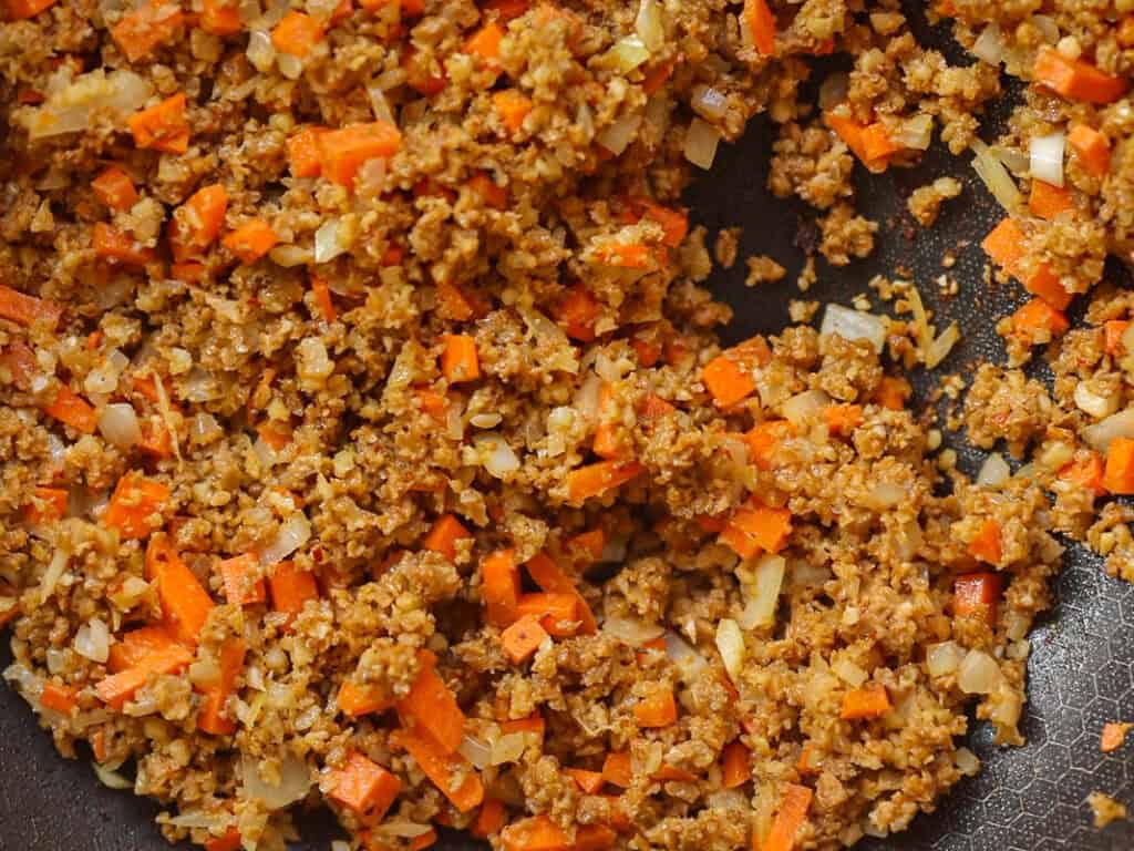 Close-up of sautéed vegetables mixed with cooked ground vegan italian sausage in a non-stick pan. The mixture includes finely chopped carrots and onions, creating a colorful and textured dish.